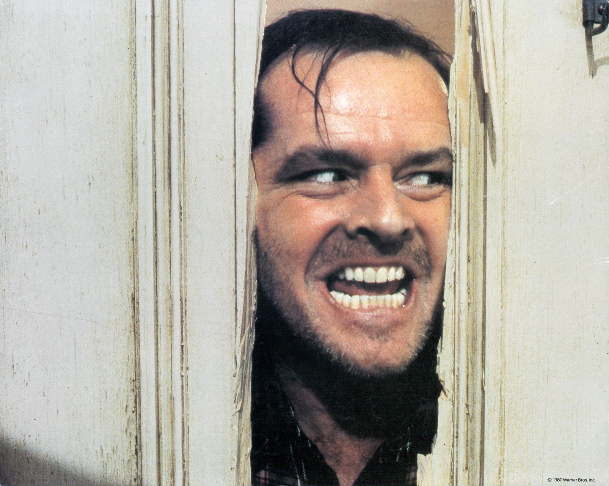 Jack peers through the splintered door while smiling in a way that feels both manic and theatrical.